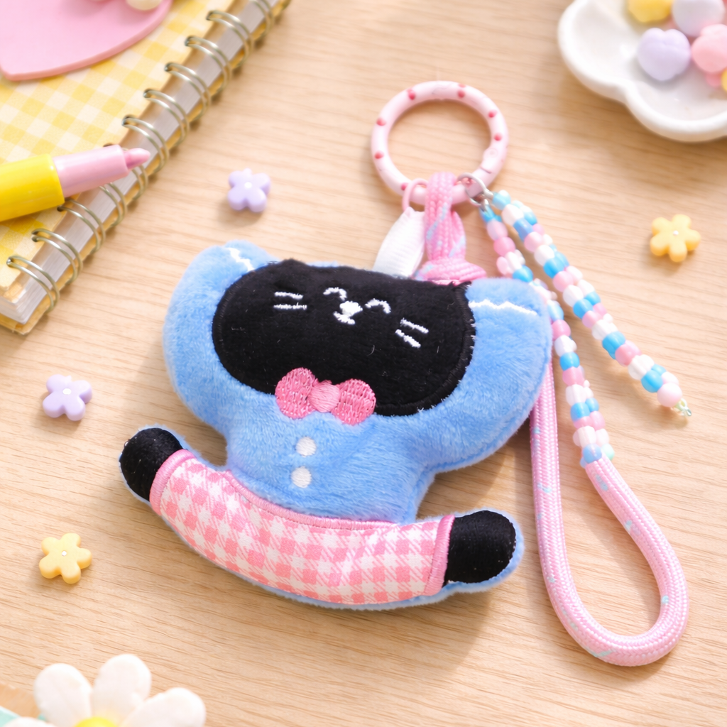 Plush keychain of a black cat with a blue dress and pink bow on a wooden surface.