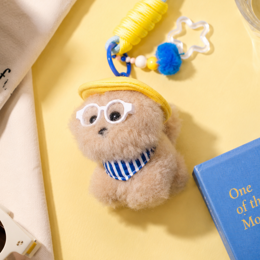 Small plush toy with a yellow hat and blue glasses on a yellow surface with a book and coffee cup.