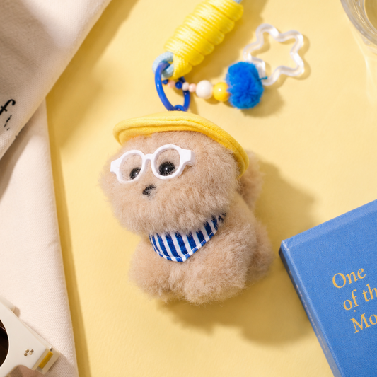Small plush toy with a yellow hat and blue glasses on a yellow surface with a book and coffee cup.
