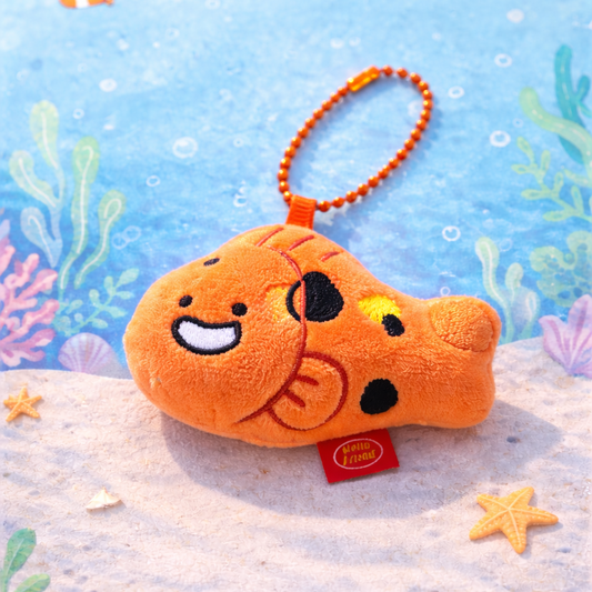 Plush fish keychain with a smiling face on a sandy beach background with sea-themed decorations.