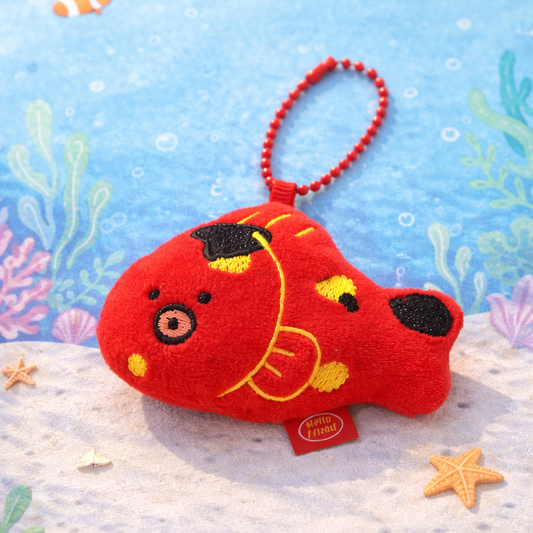 Red fish-shaped plush toy with a keychain on a sandy beach background with sea-themed decorations.