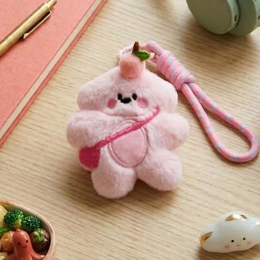 Pink plush keychain on a wooden surface with a notebook, pen, and small container.