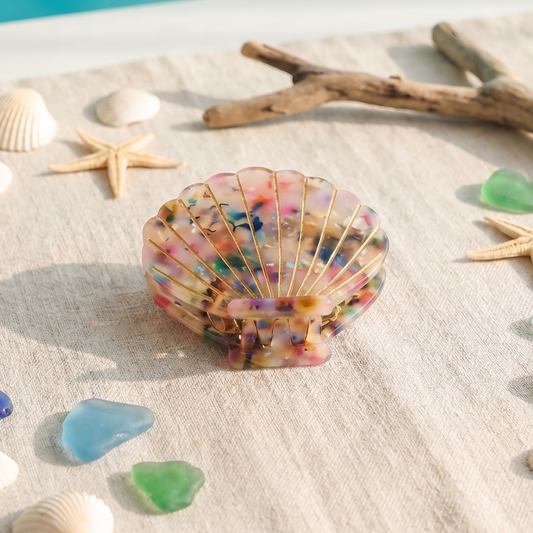 Colorful seashell hair claw on a sandy surface with seashells and starfish around, against a blue water background.